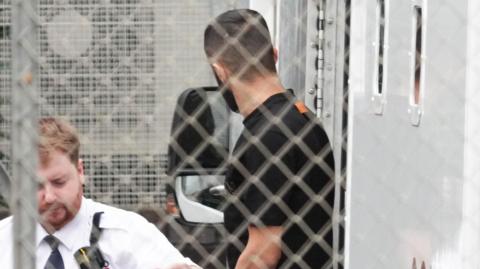 A man in a dark T‑shirt is near a van, photographed through metal fencing, with a security officer in the foreground.