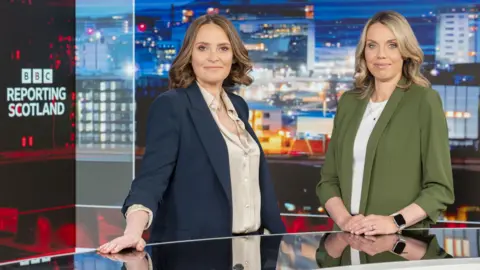 Laura Miller and Laura Goodwin standing behind the Reporting Scotland desk