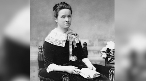 A black and white image of a woman with her hair in a plaited bun on top of her head. She is wearing early 20th-century clothes that include a large white lace collar and a black dress. She is sitting in an ornate chair, and the background is blurred.