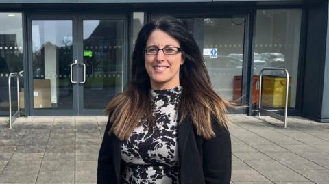 Menyw gyda gwallt hir tywyll syth a sbectol yn gwisgo siaced ddu a ffrog ddu a gwyn. Mae'n sefyll o flaen adeilad ysgol gyda'r gair 'Croeso' arno.