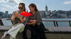 香港尖沙咀海旁兩位女士背對維多利亞港與香港島海岸線風景就坐於長椅上查看智能手機，其中一人手握一面小型中國國旗（18/3/2026）