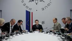 Макрон сидит во главе стола, его окружают люди в военной форме и в костюмах. За ним — флаги ЕС и Франции.