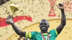 Sadi Mane in green Senegal jersey seen from chest up lifting the golder Afcon trophy. He is holding it aloft in his right hand while also lifting his left into the air. He has a red medal ribbon around his neck and golden ticker tape is seen falling in the backgroun