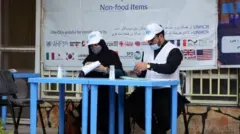 تصویر دو کارمند ملل متحد در یکی از مراکز توزیع کمکهای نقدی. این کارمندان پشت میز آبی نشته و کلاهی با نشانی ملل متحد به سر دارند. در عقب شان یک بنر ملل متحد دیده میشود.