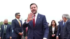 Visi Perezida w'Amerika JD Vance apepera n'ikiganza. Yambaye ikositimu y'ubururu, ishati y'umweru na karuvati itukura. Inyuma ye n'iruhande rwe hari abantu benshi. 