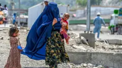 A woman carries a child in her arm. A young girl is walking alongside her. The woman is wearing an outfit in camouflage colours, and her face is covered with a long, blue veil