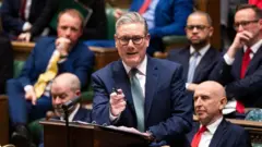 紺色のスーツと水色のネクタイを着たスターマー氏が下院の答弁台に向かい、右手のペンで前を指しながら話している。背後に、ヒーリー国防相が座り、ほかに労働党議員たちが並ぶ