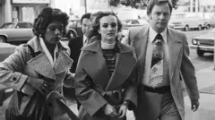 Patty Hearst, une jeune femme blonde vêtue d'un pull à col roulé et d'un trench-coat, est escortée par deux agents du FBI, une femme et un homme.