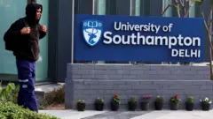 A man wearing a brown hoodie and blue trousers passes by the entrance to the campus. On his side is a board which reads University of Southampton Delhi. There are trees and shrubs on the side and a glass building in the backdrop.