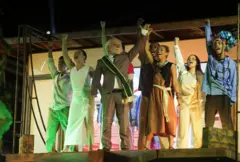 Ator com máscara de Lula, vestindo terno e faixa presidencial, ergue a mão cercado de atores que interpretam seus apoiadores