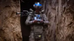 Розвідувальний дрон у повітрі між стінами окопу, на тлі в розмитті його оператор у військовій формі та екіпіруванні з синіми стрічками