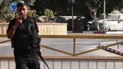 A Palestinian security officer with a gun stands in front of a lowered barrier on the only crossing between the Israeli-occupied West Bank and Jordan from 24 September 2025