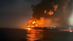 A large tanker with orange flames raging and smoke spiralling, in the night. 