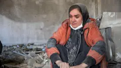 A woman sitting crying covered in dust as she sits surrounded by rubble