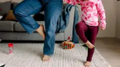 নীল জিন্সের প্যান্ট পরা একজন নারী এবং মেরুন পায়জামা ও সাদা-লাল ছোপ ছোপ টিশার্ট পরা একটি শিশু কার্পেটের ওপর এক পা তুলে দাঁড়িয়ে আছে। তাদের শরীরের ওপরের অংশ ক্যামেরার ফ্রেমের বাইরে। তাদের পেছনে একটি সোফা দেখা যাচ্ছে। কার্পেটে কয়েকটি খেলনা ও একটি ফিডার রাখা আছে 