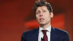 Sam Altman na czerwonym tle, ubrany w niebieski garnitur i czerwony krawat w białe kropki, patrzy w górę i w bok.