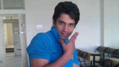 A young man wearing a blue T-shirt strikes a pose and smiles while looking at the camera in a college classroom