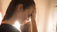 una mujer se toca la frente con la mano. Tiene rostro preocupado. Al fondo se ve la cabeza de un bebé de espaldas.