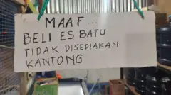 Sebuah poster tulisan manual di sebuah warung menunjukkan kepada pembeli bahwa pedagang tidak menyediakan kantong plastik untuk pembelian es batu. Poster ini bergantung dengan latar belakang plastik galon air mineral.