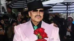 Willy Chavarria no tapete vermelho, com chapéu preto, óculos escuros e smoking rosa-claro com lapelas largas, segurando rosas vermelhas, cercado por fotógrafos
