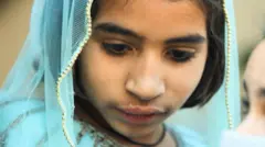 A close-up of a girl's face. She has dark hair and is wearing a blue veil loosely over the top of her head