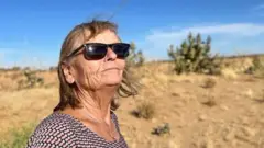 Eva Gretzmacher lève les yeux, dissimulée derrière des lunettes de soleil. Un ciel bleu et des broussailles désertiques se dessinent à l'horizon. Elle porte un collier avec un pendentif en forme de bague.