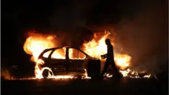 A protester walks by a burning car during clashes with police in Le Port, French Indian Ocean island of La Reunion, on June 30, 2023, three days after a 17-year-old boy was shot in the chest by police at point-blank range in Nanterre