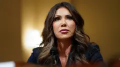 Кристи Ноэм Ички хавфсизлик бўйича Қўмита мажлисида қатнашди, у камерадан юқорига жиддий чеҳра билан қараб турибди.