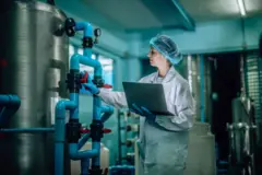 Inspektorka ds. jakości wody pitnej monitoruje systemy odwróconej osmozy. Ma na sobie biały fartuch, niebieskie rękawice, a w lewej ręce trzyma laptopa