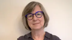 Maryellen MacDonald é uma mulher branca, de cabelos grisalhos e curtos. Ela está sorrindo para foto e usa óculos.