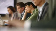 یوه ښځه چې له ورایه ستړې ښکاري، په یوه غونډه کې د خپلو همکارانو ترمنځ کمپیوټر کې کار کوي. 