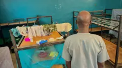 Father of one of di abducted children inside di dormitory of di school. E dey look some clothes wey dem hang for bed for di school.