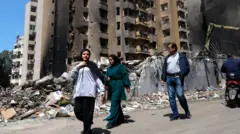 Arkada saldırıların hedefi olmuş, kısmen yıkık bir bina ve önünde yürüyen iki kadın, bir erkek. 