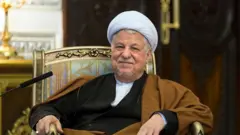 اکبر هاشمی‌رفسنجانی، با عبای قهوه‌ای روی صندلی با دسته طلایی