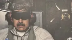 Мадуро уйги кийимида кўзлари қора тўсиқ билан қопланган, қулоқлари ҳам қулоқчин билан беркитилган ҳолда кўринишидан ҳарбий вертолёт ўриндиғида ўтирибди