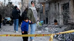 A woman with her child stands behind police tape next to rubble and debris on a street and looks at the aftermath of an Israeli and the U.S. strike on a police station, 2 March 2026.