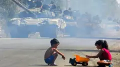 جاده خاکی با ردیف تانک‌ها و دو کودک در کنار جاده در حال بازی با یک کامیون اسباب‌بازی