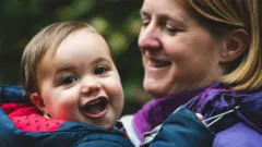 James, yang masih bayi sedang digendong ibunya Laura di pelataran dengan latar belakang peohonan hijau.