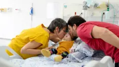 Foto de um bebê em uma cama de hospital com um homem beijando sua bochecha de um lado, e uma mulher, do outro.