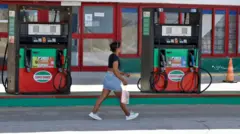 デニムのショートスカートに黒い半袖トップスを着た女性が、ガソリンスタンドの前を歩いている。ガソリンスタンドの窓ガラスには、白地に黒い文字で「cerrado」（スペイン語で「閉鎖」）と書かれたサインが掲げられている。赤と緑の固定式計量機械のデジタル表示は消えており、稼働していないことがわかる