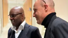 Charles Onana ari kumwe na Damien Sirieix, bamwenyura. Barahagaze, bameze nk'abarimo gutambuka, Onana yambaye indorerwamo, naho Sirieix we yazamuye indorerwamo hejuru ku mutwe