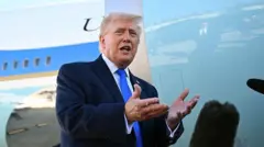 大統領専用機を背に、スーツに青いネクタイを着けたトランプが、複数のマイクの前で手振りを交えている