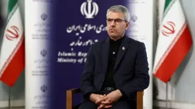 Umuvugizi wa minisiteri y'ububanyi n'amahanga ya Irani Esmail Baghaei yicaye ku ntebe ndende yegamirwa, areba muri 'camera'. Yambaye ikositimu y'umukara, inyuma ye hari amabendera abiri ya Irani.