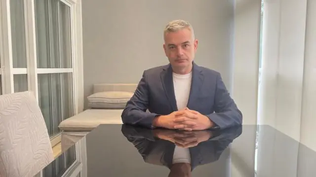 Батько Вікторії сидить за скляним столом, склавши руки разом. У нього сиве волосся, він у сірому піджаку та білій футболці.