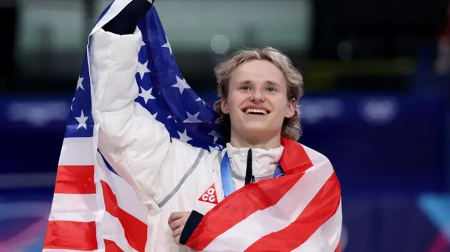 Укутанный в американский флаг Малинин машет рукой на церемонии награждения