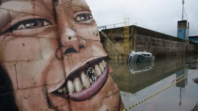 Um grande mural de rosto humano ocupa um muro de concreto, com destaque para o seu sorriso. Ao lado, uma área está alagada e um carro aparece parcialmente submerso na água. O ambiente é cinzento.