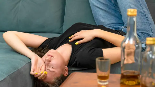 Une jeune femme allongée à l'envers sur un canapé, les yeux fermés et la main sur le visage, avec des bouteilles presque vides posées sur la table devant elle.