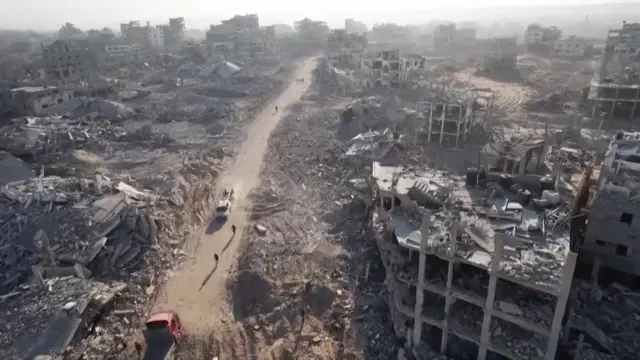 The view from a drone flying above Gaza City shows a decimated landscape. A road with two vehicles and people on it is surrounded by flattened buildings. One building to the right has three storeys with the walls blown out. Most of the other buildings are in ruins.