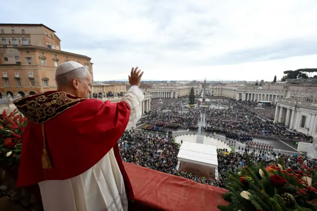 Pope Leo, wearing white and red papal vestments, waves at crowds from the balcony of St Peter’s Basilica on Christmas Day 2025.