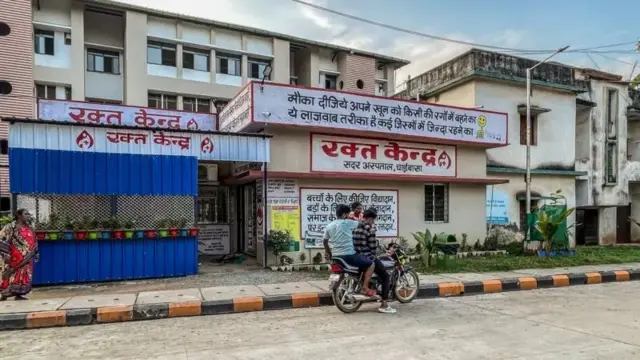 একটি ভবন যেখানে ব্লাড ব্যাংক রয়েছে, এর সামনে লাল রংয়ে হিন্দিতে কিছু লেখা। ভবনটির সামনে মোটরসাইকেলে বসে আসেন দুই তরুণ, পাশে দাঁড়িয়ে আছেন একজন নারী। একটু দূরে লাল শাড়ি পরা আরো একজন নারী দাঁড়িয়ে আছেন।